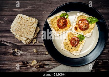 Rice crackers with chickpea hummus, cherry tomato and fresh basil on a plate and crackers on a rustic table. Healthy food. Stock Photo