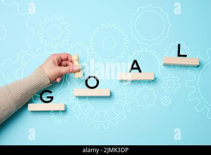 A female hand holds a wooden figurine on a blue background and wooden blocks. The concept of achieving the goal, the way forward Stock Photo