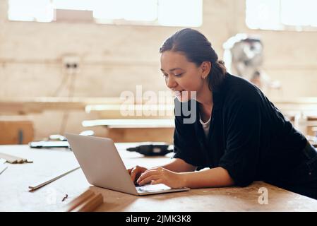 Technology has a place everywhere, even in the workshop. a young female carpenter using a laptop while working inside her workshop. Stock Photo