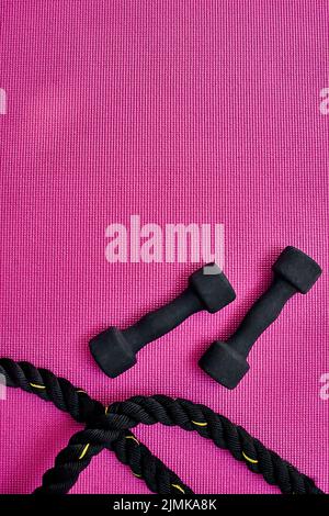 Who wants to get fit. High angle shot of two lightweight dumbbells and piece of rope placed on a pink background inside of a studio. Stock Photo