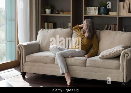 Happy beautiful grey haired elderly woman sitting on comfortable couch Stock Photo