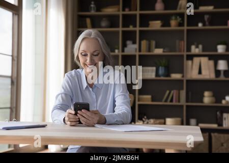 Happy older retired lady using online app smartphone at home Stock Photo