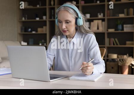 Senior mature student woman in big headphones watching learning webinar Stock Photo