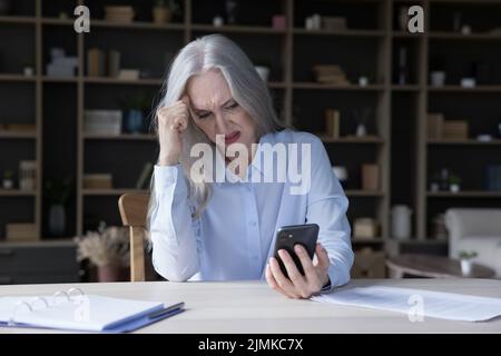 Frustrated annoyed elderly retired woman holding wrong working mobile phone Stock Photo