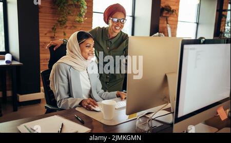 Two Muslim businesswomen having a discussion while looking at a computer screen in an office. Cheerful businesswomen working as a team in a modern co- Stock Photo