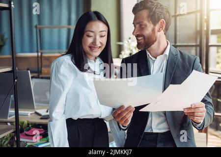Two colleagues looking on documents and smile standing in office Stock Photo