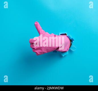 Hand in a pink household rubber glove sticks out of the torn hole and shows a gesture okay, thumb up. Blue background Stock Photo