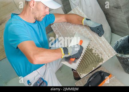 Professional Tile Installer Applying Adhesive to the Tile Before Installing It in His Client’s Bathroom. Bathroom Remodeling Work. Side View. Stock Photo