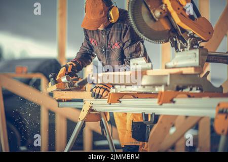 Carpentry Construction Worker Planing the Wood Using Electric Planer After Adjusting the Plank with Circular Saw. Wooden House Building Site Theme. Stock Photo