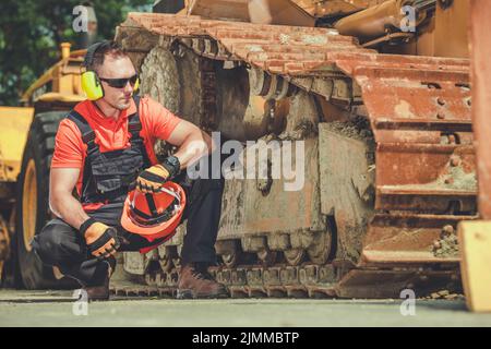 Caucasian Construction Worker Wearing Noise Reduction Headphones and Holding Red Hard Hat in His Hand Resting in Front of His Heavy Industrial Equipme Stock Photo