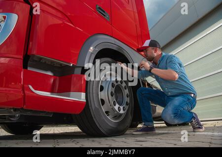 Caucasian Trucker Closely Checking the Condition of the Tires in His Large Heavy Duty Red Semi Truck Before Setting Off to Deliver a Cargo. Transporta Stock Photo