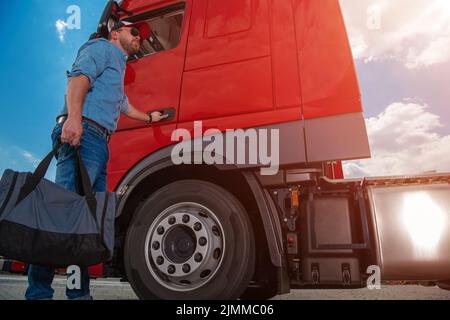 Caucasian Lorry Driver With a Bag in His Hand Opening the Cab Door to His Red Semi Truck Ready to Start Another Route and Complete a Delivery. Heavy D Stock Photo