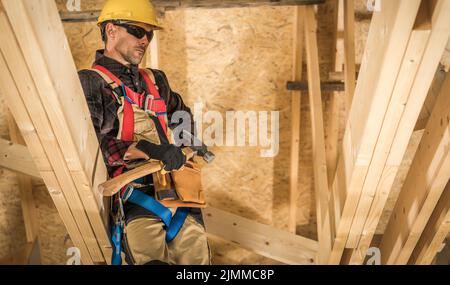 Caucasian Middle Aged Builder in Yellow Helmet, Work Uniform and Safety Harness Standing in the Middle of Wooden Residential House Construction Site w Stock Photo