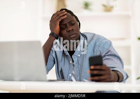 Bored Black Freelancer Guy Sitting At Desk And Holding Smartphone In Hand Stock Photo