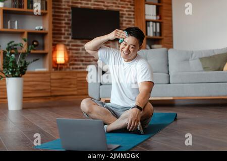 Happy asian middle aged man wiping sweat after home workout, sitting on yoga mat and looking at laptop Stock Photo
