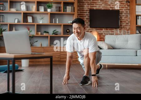 Home training. Happy asian mature man doing stretching exercises for legs in front of laptop, watching online tutorials Stock Photo