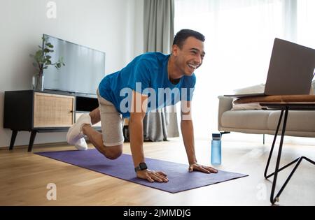 Active arab man in sportswear exercising at home, working on abs and arms strength, looking at laptop screen Stock Photo