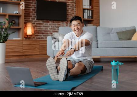 Home training concept. Positive korean man stretching leg muscles in front of laptop, watching video tutorials online Stock Photo