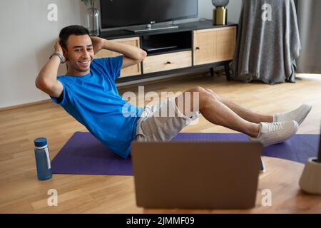 Happy arab man working on abs, using laptop and fitness mat at home, exercising in living room Stock Photo