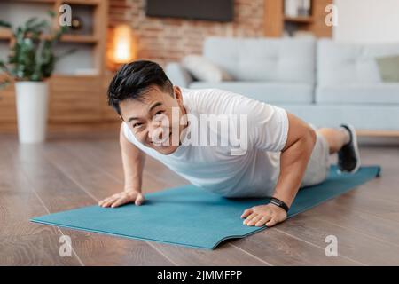 Strong asian mature man making strength workout, standing in plank pose or doing push ups at home on yoga mat Stock Photo