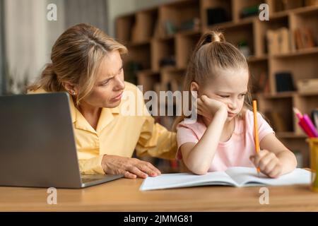 Worried mother looking at offended daughter while doing homework together, sitting at desk with laptop and notebook Stock Photo