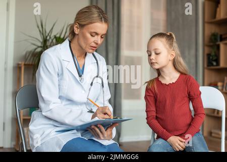 Focused female doctor writing down girl's symptoms on clipboard while child looking what doc writing in chart Stock Photo