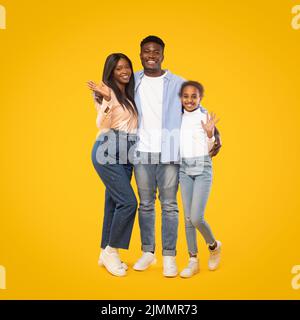 Portrait of happy african american family of man, woman and girl waving hands while posing over yellow background Stock Photo