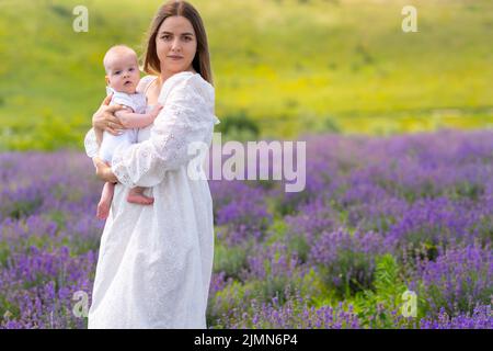 Young mother standing in a lavender field holding a little baby in her arms as they both look at the camera Stock Photo