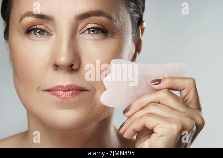 Beautiful middle aged woman making facial massage with a rose Gua Sha stone Stock Photo