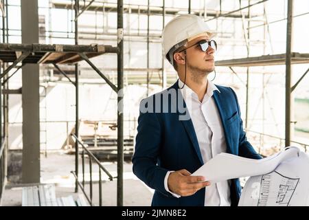 Architect wearing formal suit and hard hat holding a blueprints on a construction site Stock Photo