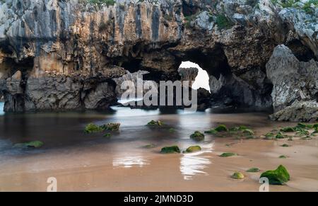 Beach with cliffs and caves and algae covered rocks on golden sand Stock Photo