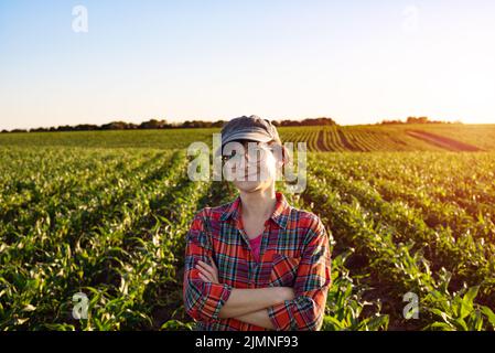 Middle age female caucasian confident satisfied smiling farm worker with crossed arms stands at corn field Stock Photo