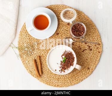 Yogurt with chocolate granola in cup, breakfast with tea on beige background, top view. Stock Photo