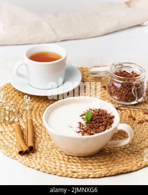 Yogurt with chocolate granola in cup, breakfast with tea on beige background, side view, vertical. Stock Photo