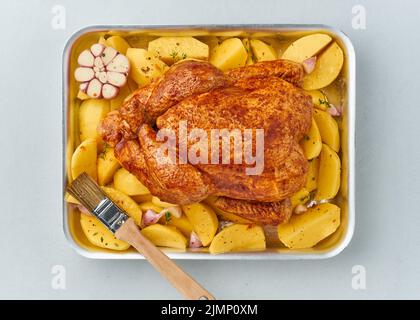 Whole marinated chicken in sauce with raw potatoes, with brush in baking dish, top view. Stock Photo