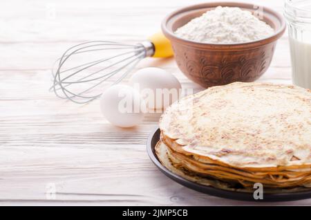 Stack of French crepes in frying pan on wooden kitchen table with milk eggs and flour aside Stock Photo