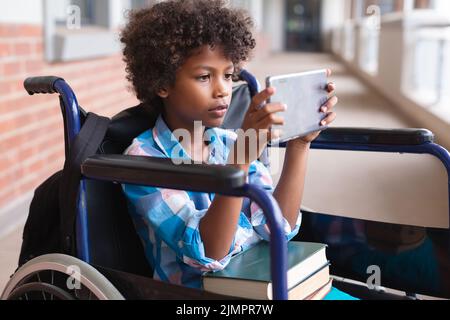 African american elementary schoolboy using digital tablet while sitting on wheelchair in corridor Stock Photo