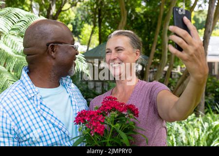 Happy multiracial senior couple with flowers taking selfie through smart phone in backyard Stock Photo