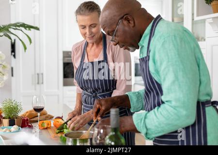 Senior multiracial couple in aprons preparing food together in kitchen at home Stock Photo