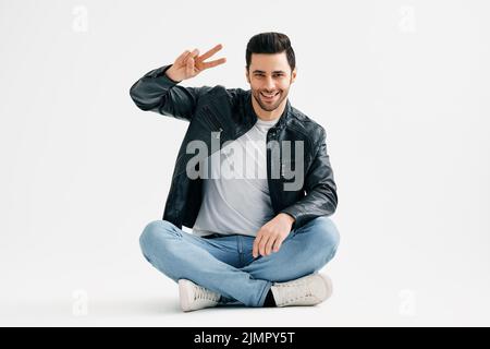 Positive cheerful young man doing victory sign sitting with legs crossed on floor isolated over white background Stock Photo