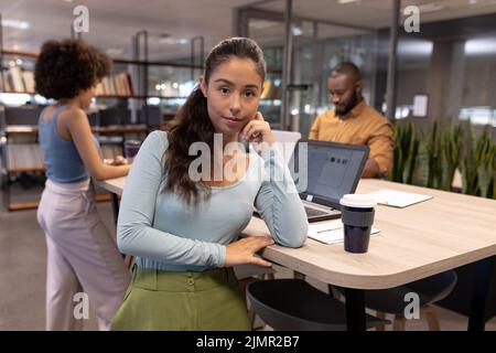 Portrait of confident young hispanic businesswoman leaning on desk against biracial colleagues Stock Photo