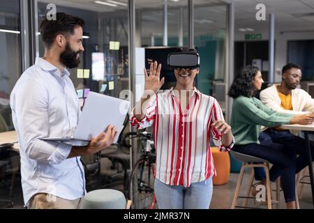 Biracial businesswoman wearing vr headset by caucasian businessman with laptop during meeting Stock Photo