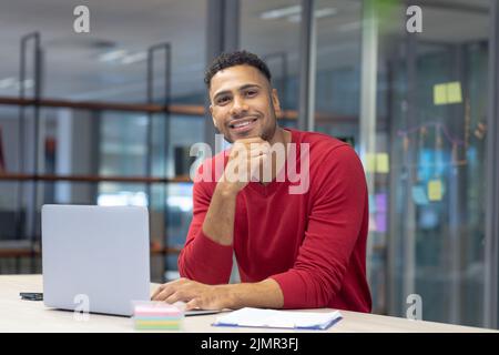 Portrait of happy hispanic male advisor sitting with laptop and document at modern workplace Stock Photo