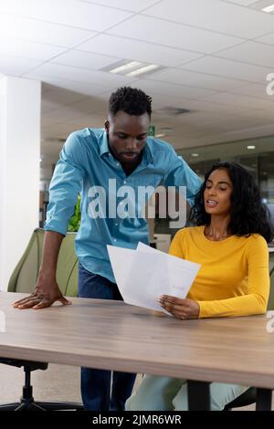 Biracial male and female colleagues discussing over document at desk in ...