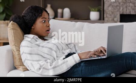 African girl student freelancer user lies on sofa at home looking at laptop using internet checks mail reads unexpected bad news feels shock surprise Stock Photo