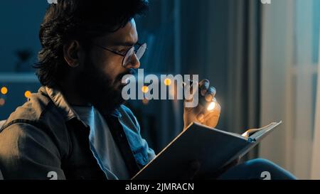 Bearded adult man in glasses at night home living room with flashlight shines on book reads preparing for exams reading bible, faith concept. 30s guy Stock Photo