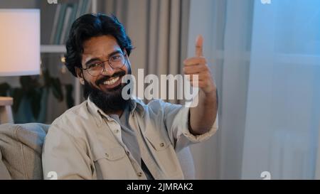 Portrait happy satisfied joyful arabic indian man bearded male in glasses sitting at home in new apartment on comfortable sofa looking at camera Stock Photo