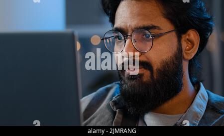 Close-up bearded male face reflection light from laptop monitor in glasses. Busy serious focused Arab Indian man looking at computer working freelance Stock Photo