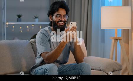 Happy arabian indian male millennial man holding modern smartphone texting message at home sitting on couch in evening bearded guy in glasses using Stock Photo