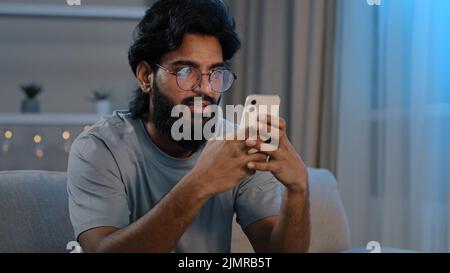 Serious busy Arabian Indian man bearded male in glasses typing browsing in smartphone texting chatting using mobile app messaging at home sitting on Stock Photo